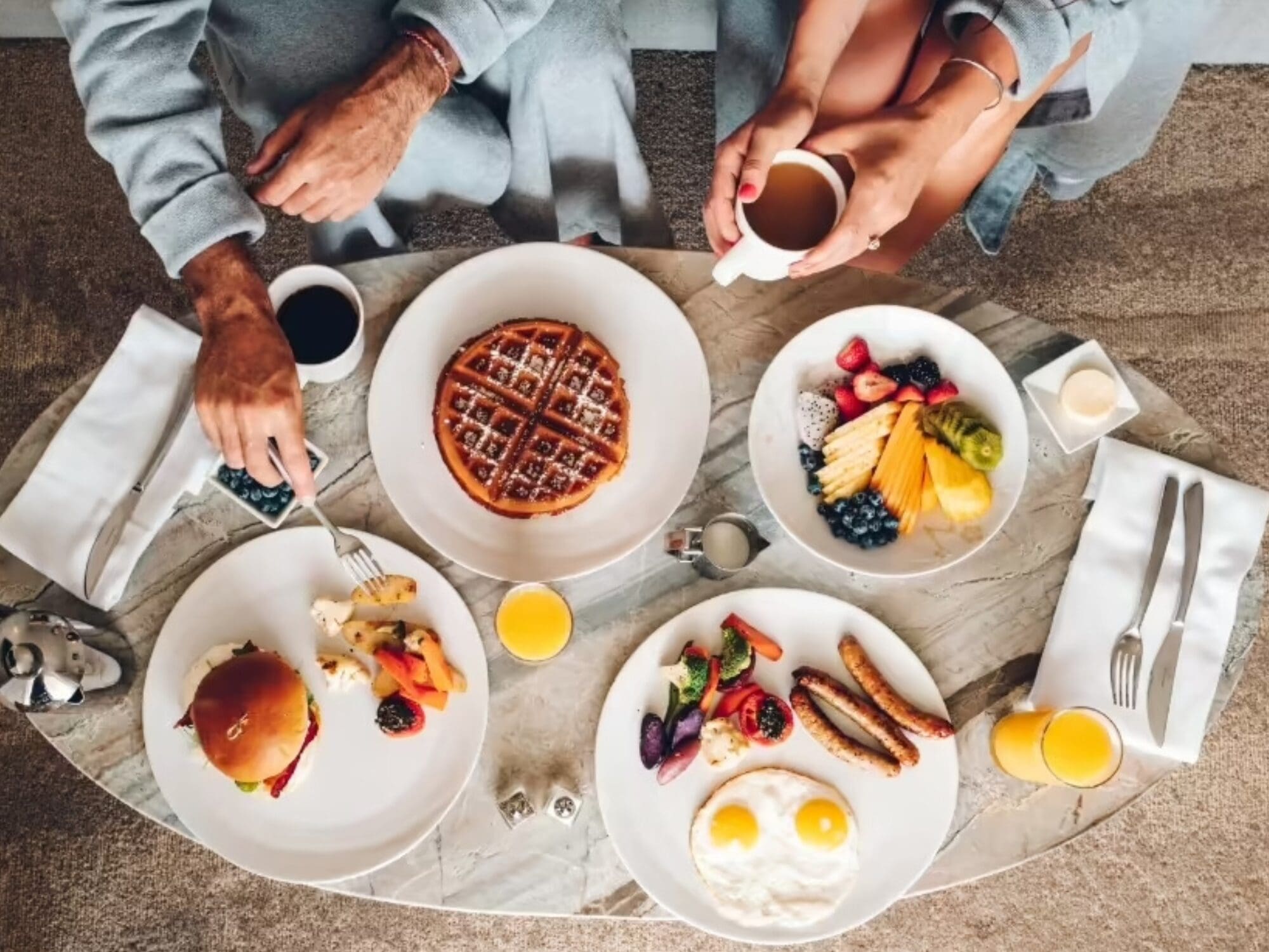 A top view of a breakfast spread on a table, featuring a waffle, plates of eggs with sausages, a burger, fresh fruit, orange juice, and two cups of coffee. Two people are sitting, one holding a cup of coffee.