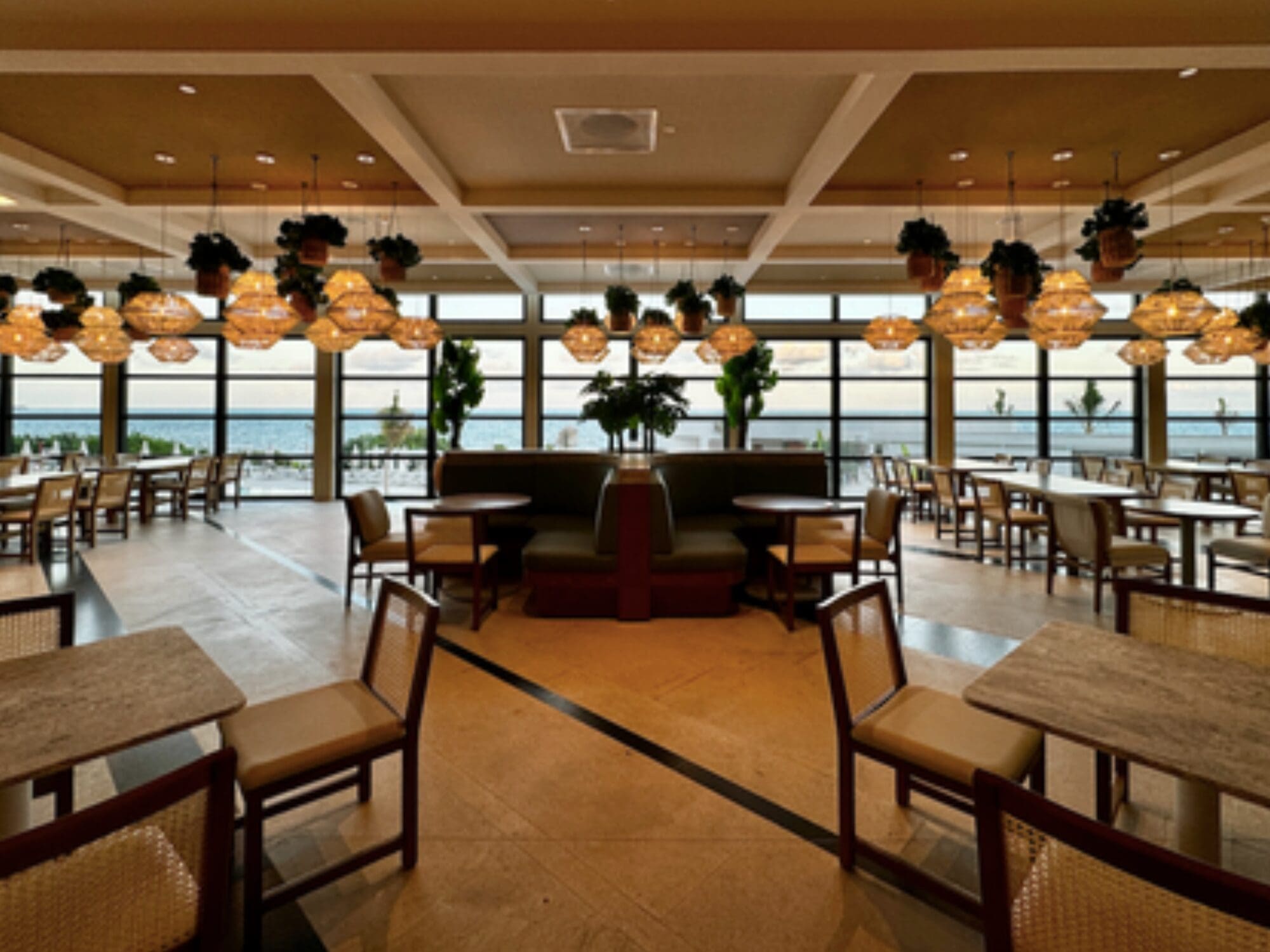 A spacious, modern dining area with wooden tables and chairs is shown. Large windows offer a view of the ocean. Hanging pendant lights and potted plants add to the decor. Natural light fills the space.