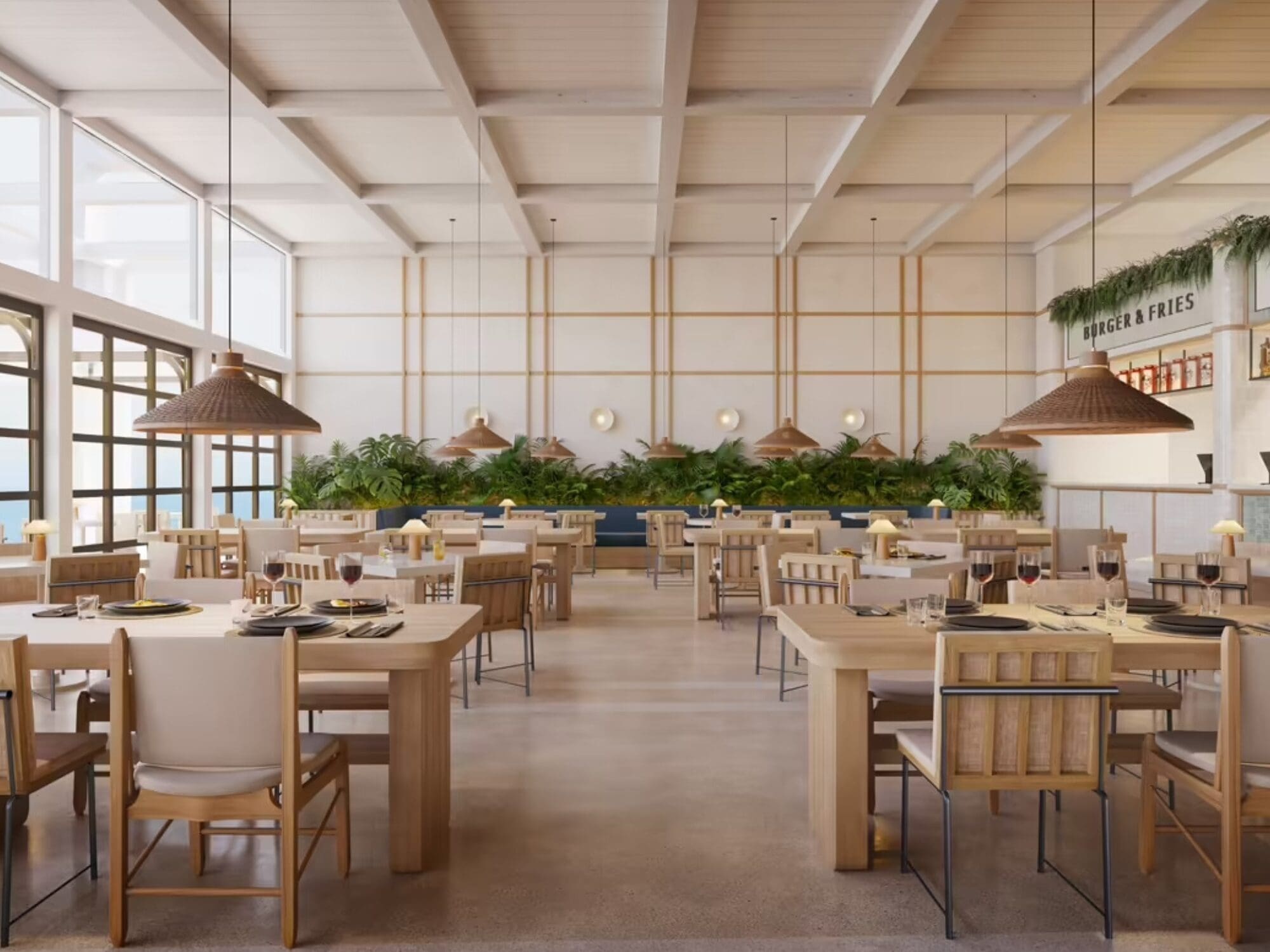 A spacious, modern restaurant with wooden tables and chairs. Large windows on the left let in natural light. Pendant lights hang from the ceiling. Green plants decorate the back wall. Tables are set with plates and glasses.