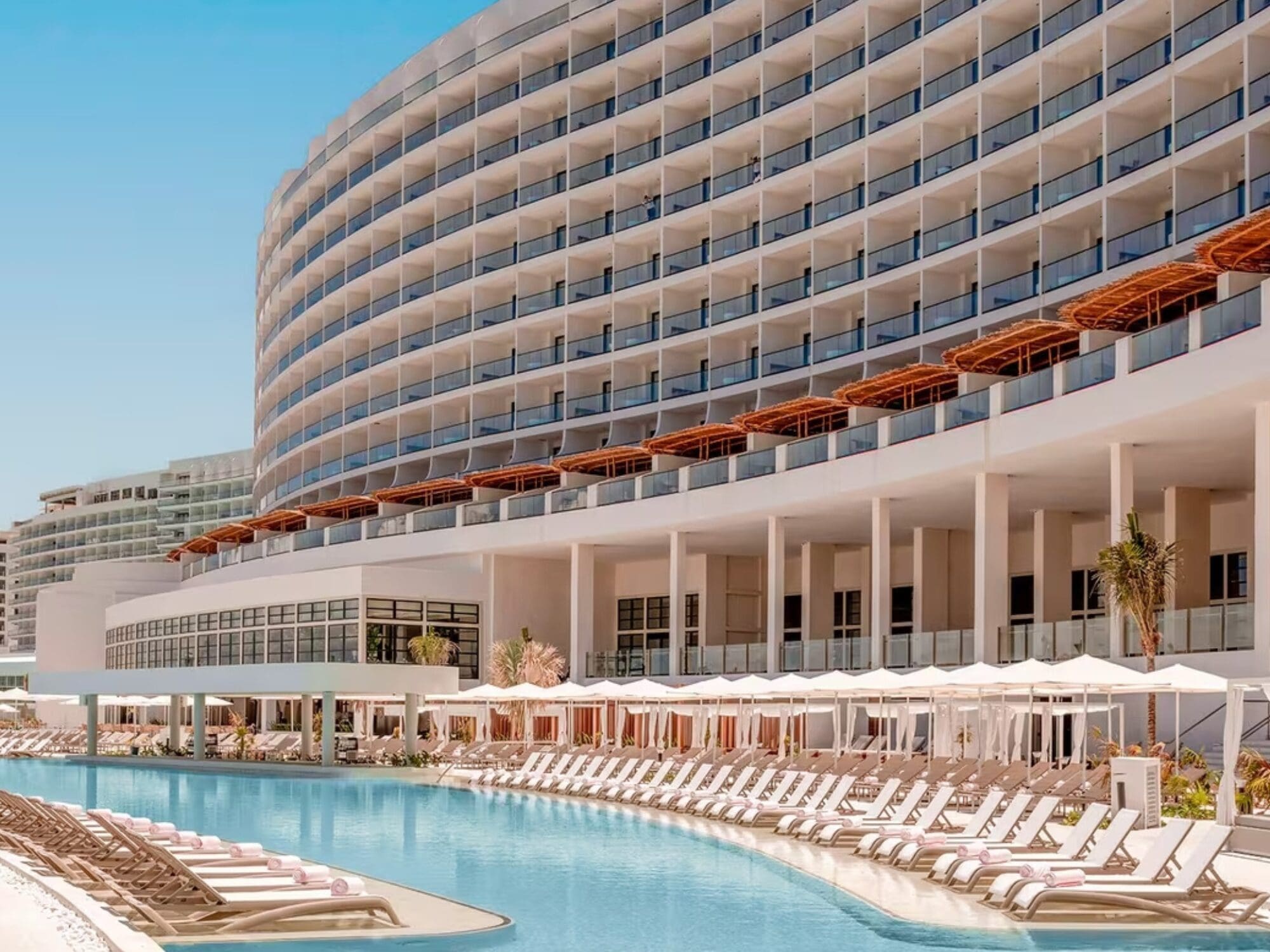 Modern resort with a curved, multi-story building featuring balconies. A large pool lined with white lounge chairs and umbrellas is in the foreground. The sky is clear and blue, creating a relaxing and luxurious atmosphere.