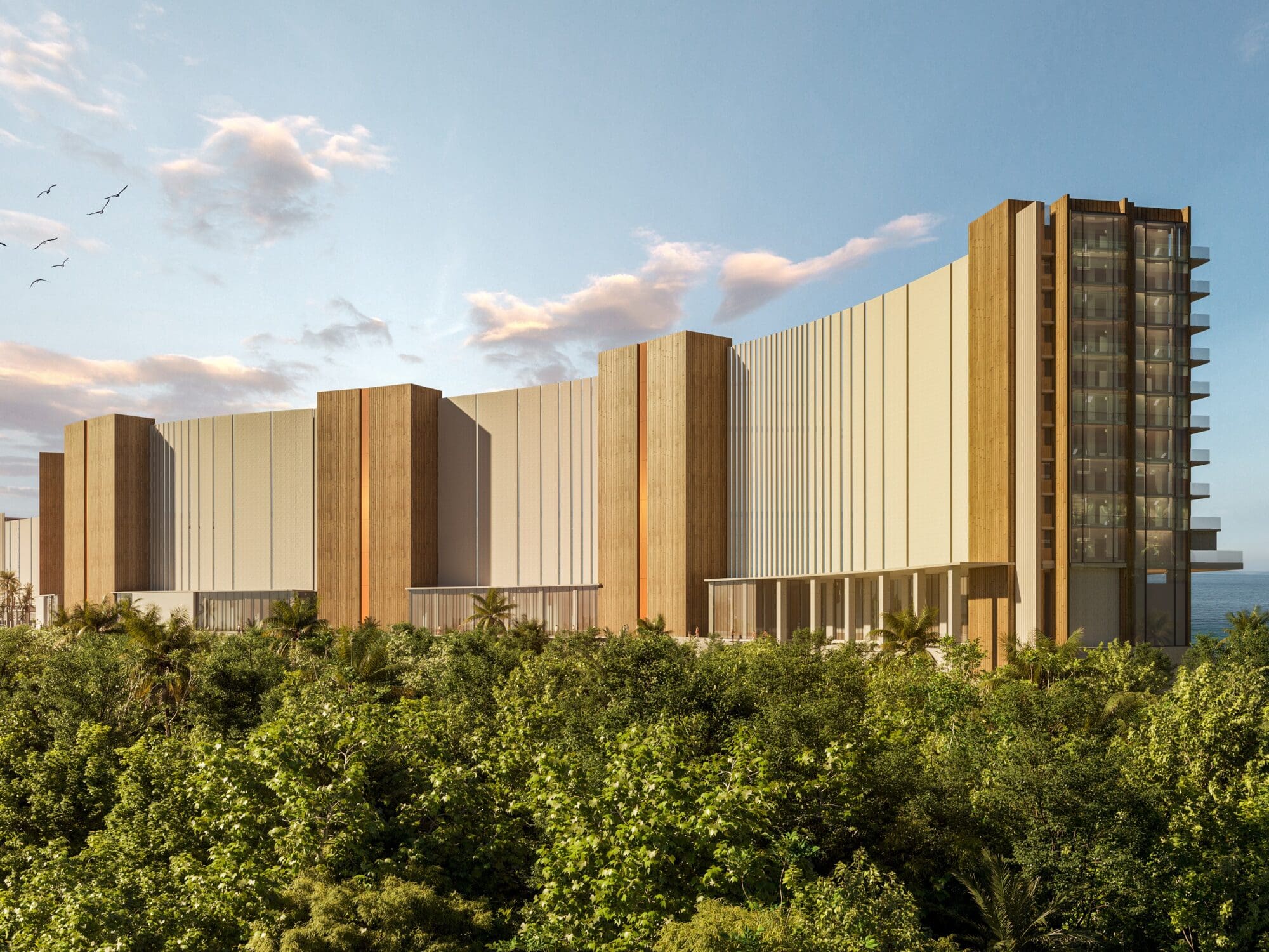 A modern building with a curving design, featuring alternating vertical sections of glass and brown panels. It is surrounded by lush greenery and set against a blue sky with scattered clouds and birds in the distance.