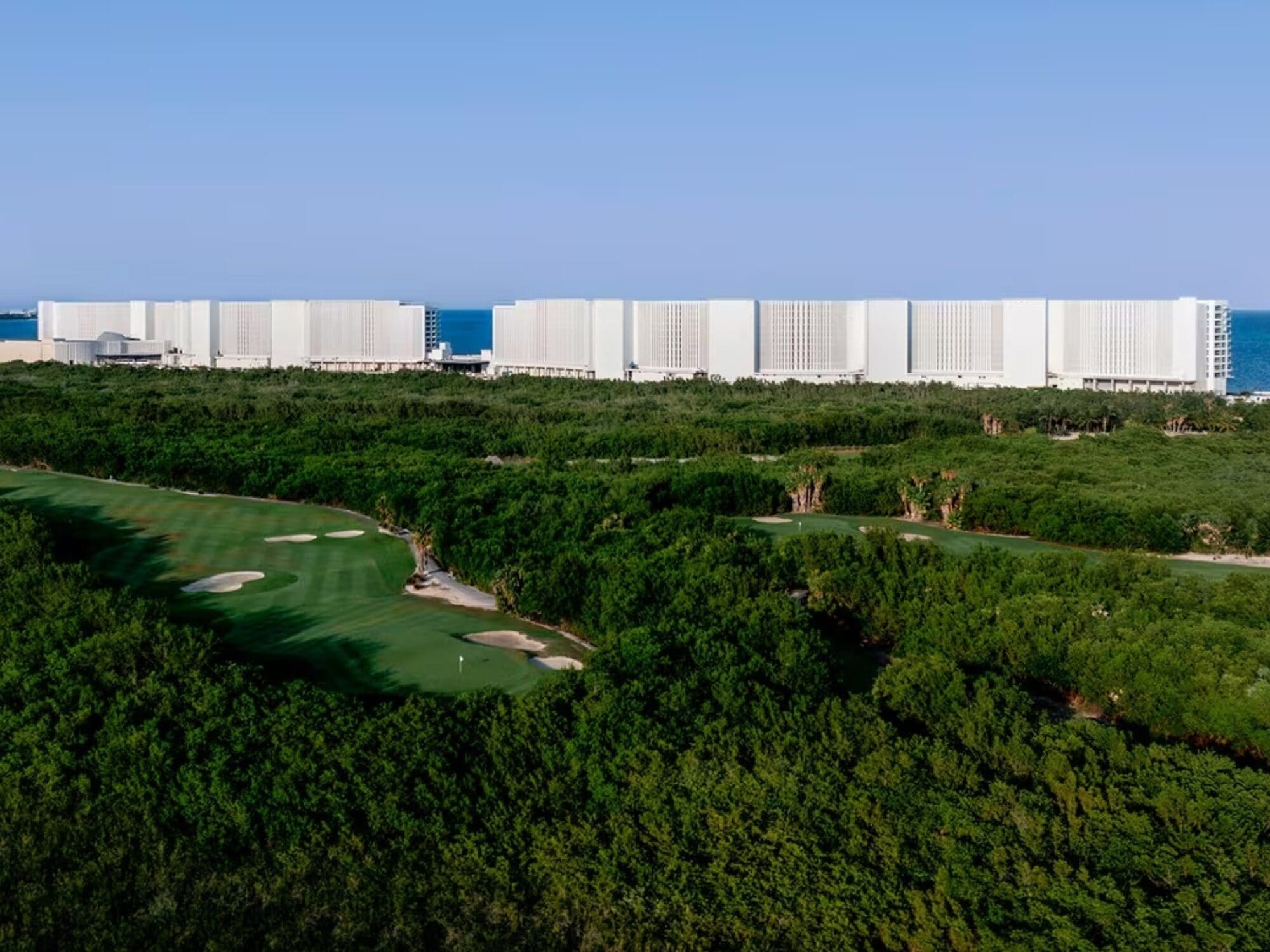 Aerial view of a coastal resort with modern white buildings near the ocean. In the foreground, a lush green golf course with several sand traps is visible, surrounded by dense forest under a clear blue sky.