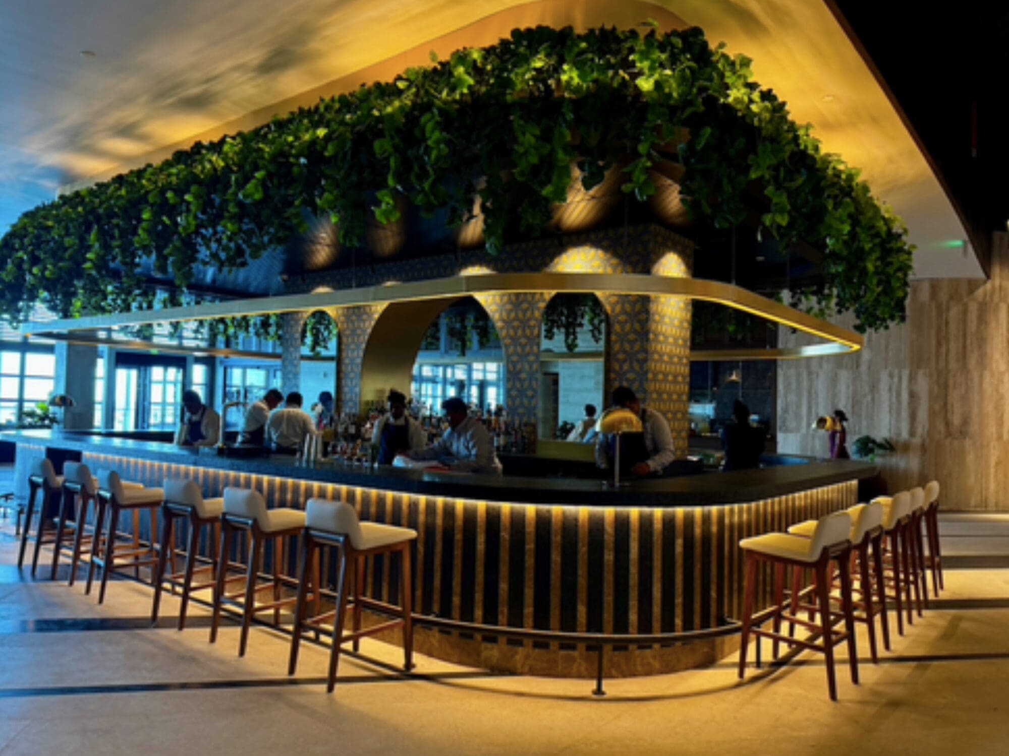Modern indoor bar with lush greenery hanging from the ceiling, a curved counter with backlit panels, high stools, and people casually seated. Soft, ambient lighting creates a warm and inviting atmosphere.