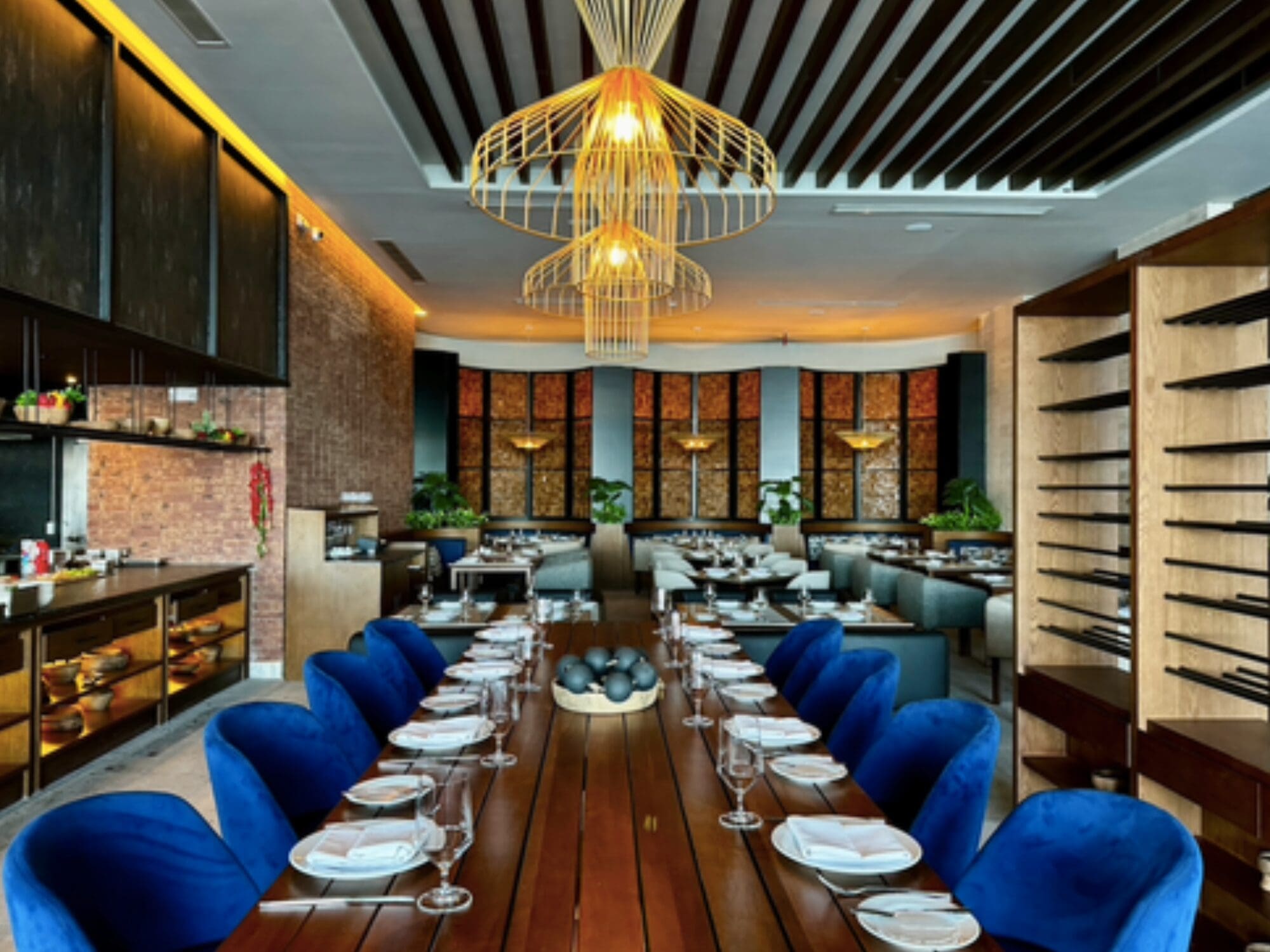 Luxurious restaurant interior with a long wooden table set with white plates and glassware. Blue velvet chairs surround the table. Modern, gold pendant lights hang from the ceiling. Brick walls and wood shelving add warmth to the decor.