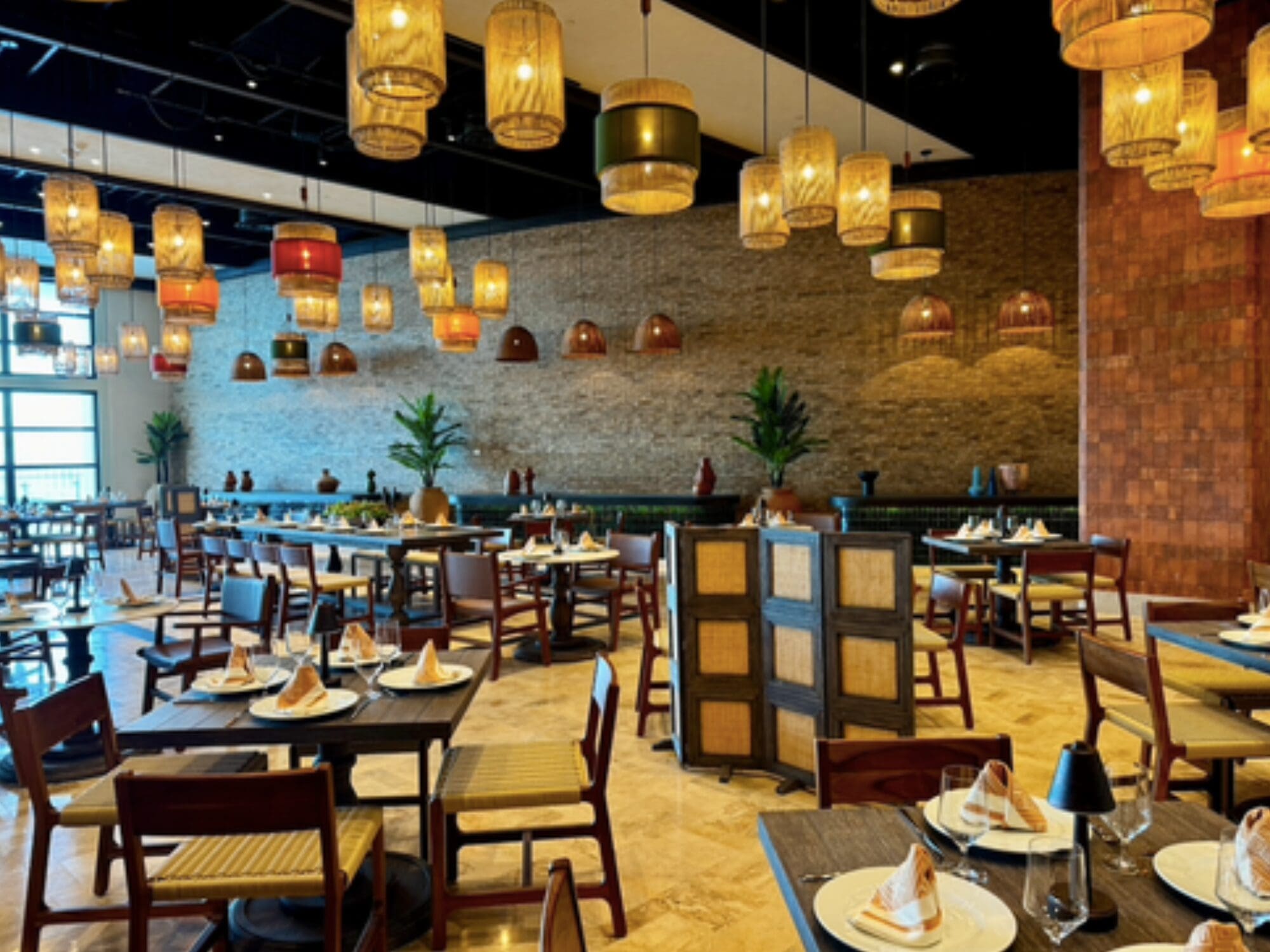 A spacious restaurant with wooden tables and chairs, set with folded napkins and plates. Warm, decorative hanging lights create a cozy atmosphere. Plants accent the room, and a stone wall adds texture in the background.