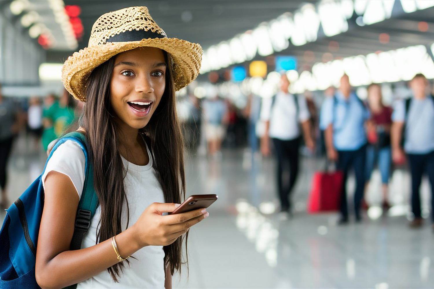 A young woman wearing a straw hat and backpack smiles while holding her phone in an airport terminal. Blurred travelers and bright lights are visible in the background.
