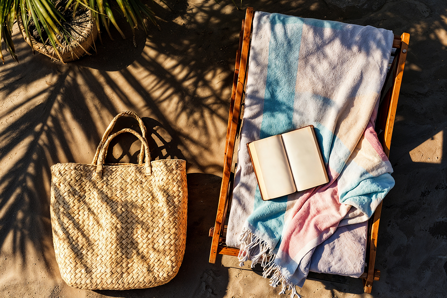 A woven beach bag sits on sandy ground next to a wooden lounge chair covered with a pastel-striped towel and an open blank notebook, with palm shadows cast across the scene.